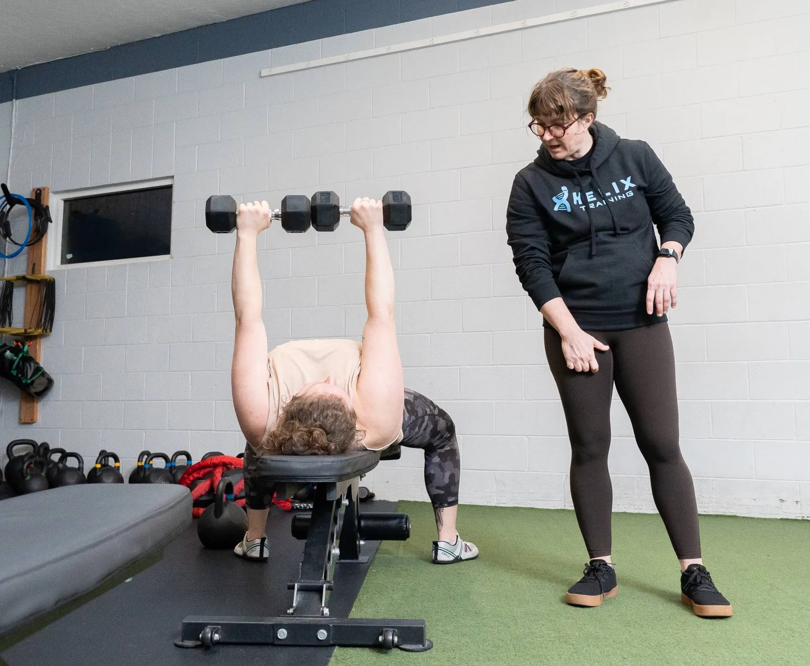 A Helix member presses dumbbells on a bench while a coach watches her form.