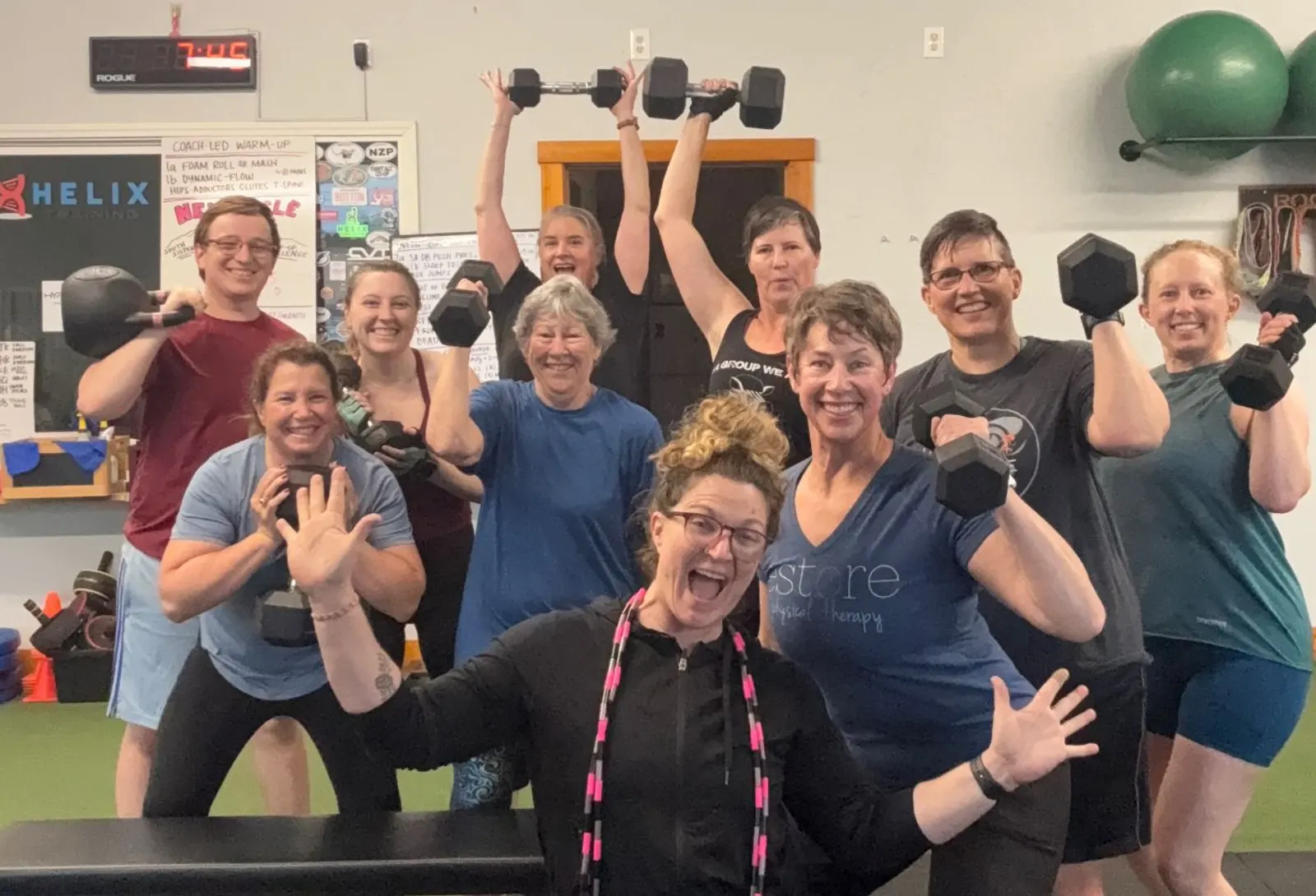 A smiling group of Helix members holding dumbbells and a kettlebell overhead and at their shoulders, with a coach in front cheering.