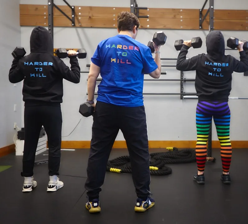 Three Helix members, seen from behind, press dumbbells overhead wearing the Helix mantra, "Harder to Kill," on their shirts and hoodies.