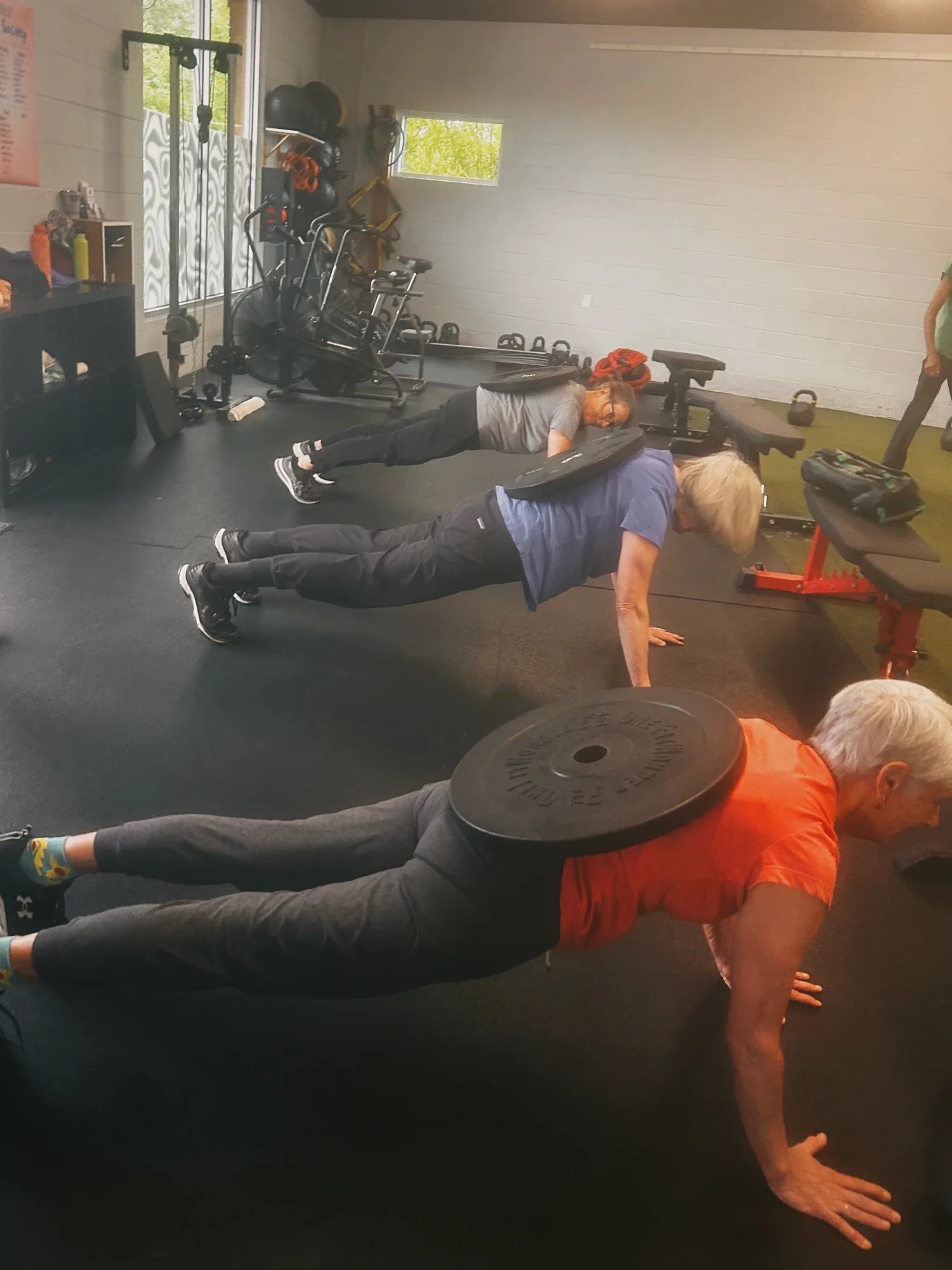 Three Helix members with gray and white hair hold plank positions on the gym floor, each with a weight plate balanced on their back.