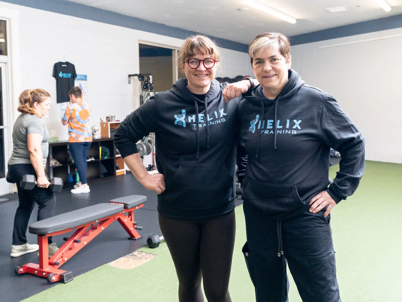 Coaches Rebecka Weinsteiger and Kathy Lynch smile side by side in Helix hoodies while members train behind them.